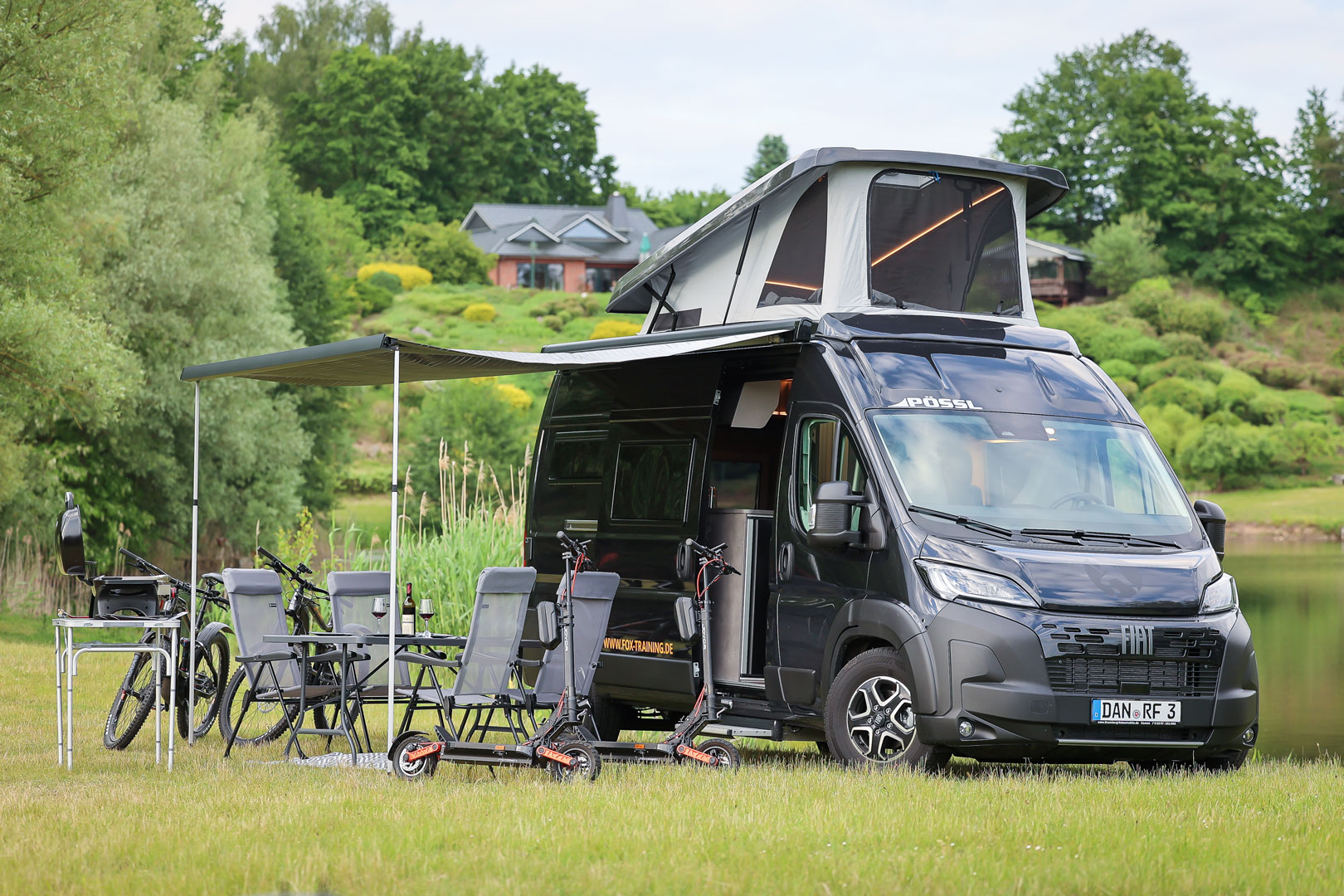 Pössl Campervan mit Aufstelldach, Markise, Campingmöbeln, E-Scootern und E-Bikes auf einer Wiese vor einem See